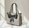 Gray handbag with black bow, double handles, sitting on a white surface in natural light